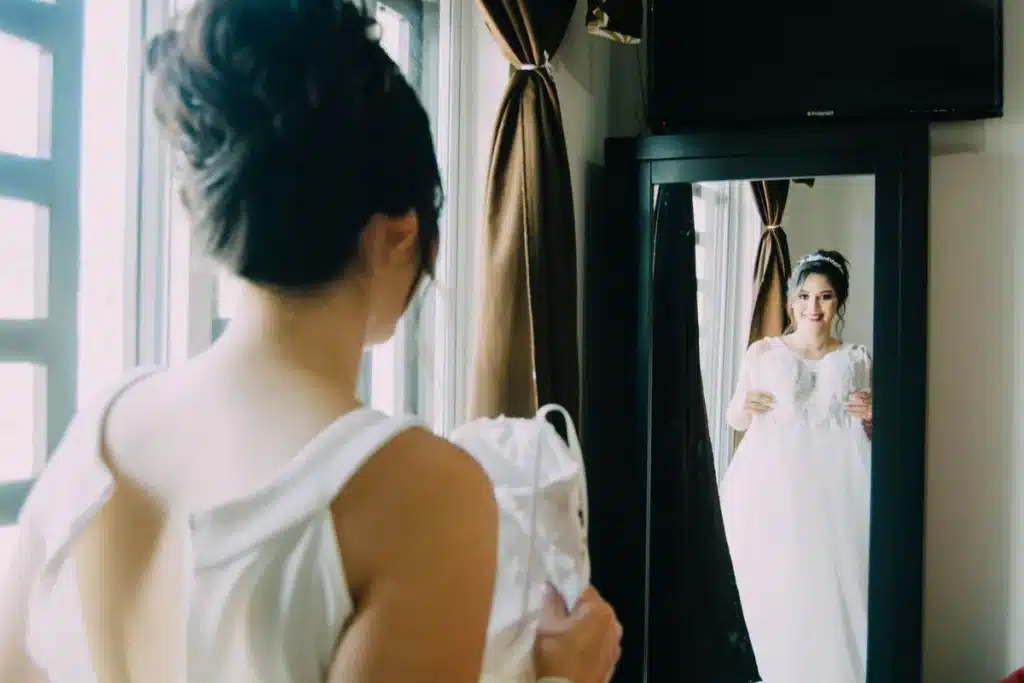 Femme ajustant sa tenue de mariage devant un miroir dans une lumière naturelle douce.
